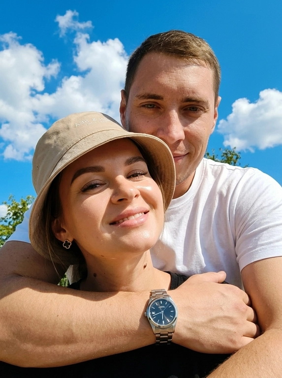 Кинематографичный кадр крупным планом (selfie style), пара в объятиях (классическое "обнимашки сзади") на фоне ярко-голубого неба с облаками.  Женщина на ней бежевая панама с брендовой надписью. Её кожа выглядит сияющей, подсвеченной мягким естественным солнечным светом. Выражение лица спокойное, глубокий взгляд, направленный прямо в камеру. Мужчина стоит строго позади девушки и обнимает её сзади, одна его рука обнимает её за плечи/шею спереди — предплечье лежит поперёк её ключиц, кисть свободно свисает с её плеча, на запястье классические серебряные часы с черным циферблатом. Мужчина в белой футболке, выражение лица доброжелательное, его взгляд прямо в объектив. Ракурс сверху, камера сфокусирована на лице девушки. Высокий контраст, насыщенные цвета, мягкая прорисовка деталей кожи. Эффект пленочной фотографии, легкое зерно, атмосфера летнего дня, эстетика социальных сетей. Телефона не видно в кадре.