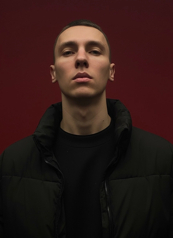 Создай портрет, используя те же черты лица (на основе загруженного фото), характеризующийся резким кинематографическим освещением и интенсивным контрастом. Снятый с немного нижнего ракурса, направленного вверх, что подчеркивает линию подбородка и шеи модели, этот портрет производит впечатление доминантности и скульптурной элегантности. Фон — глубокий, насыщенный малиново-красный, создающий яркий визуальный контраст со сияющей кожей модели и темной одеждой.