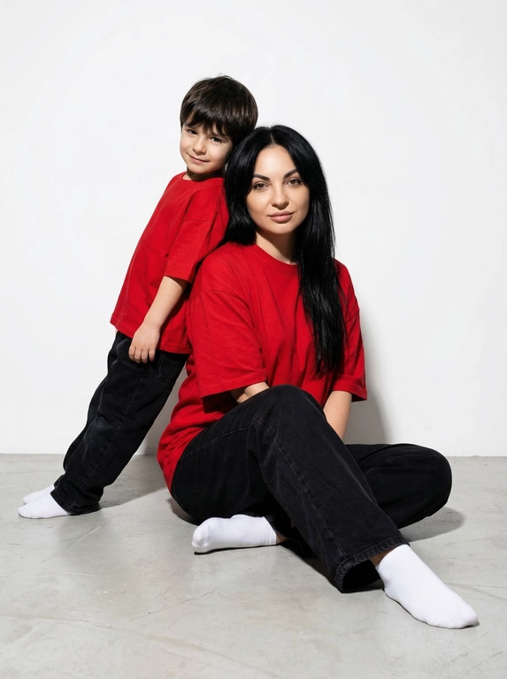 Гиперреалистичная фотография. Качество 8К. Используя портретное сходство с людьми с фотографий, создай фотографию: женщина в красной оверсайз свободной футболке и свободных черных джинсах, белых носках. Ребенок одет в красную оверсайз футболку, черные свободные джинсы и белые носочки. Женщина сидит на полу, согнув колени, смотрит прямо в кадр с еле заметной улыбкой. Сзади женщины в такой же позе сидит ребенок облокотившись спиной к спине женщины, с легкой улыбкой. Темный стиль модного редакционного журнала, глубокие тени и драматическое студийное освещение, белый фон, элегантная и мощная композиция, ультрареалистичный, высокая детализация, художественный портрет.