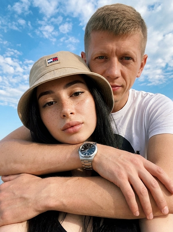 Кинематографичный кадр крупным планом (selfie style), пара в объятиях (классическое "обнимашки сзади") на фоне ярко-голубого неба с облаками.  Женщина на ней бежевая панама с брендовой надписью. Её кожа выглядит сияющей, подсвеченной мягким естественным солнечным светом. Выражение лица спокойное, глубокий взгляд, направленный прямо в камеру. Мужчина стоит строго позади девушки и обнимает её сзади, одна его рука обнимает её за плечи/шею спереди — предплечье лежит поперёк её ключиц, кисть свободно свисает с её плеча, на запястье классические серебряные часы с черным циферблатом. Мужчина в белой футболке, выражение лица доброжелательное, его взгляд прямо в объектив. Ракурс сверху, камера сфокусирована на лице девушки. Высокий контраст, насыщенные цвета, мягкая прорисовка деталей кожи. Эффект пленочной фотографии, легкое зерно, атмосфера летнего дня, эстетика социальных сетей. Телефона не видно в кадре.

Доп. характеристики: не менять черты лица парня