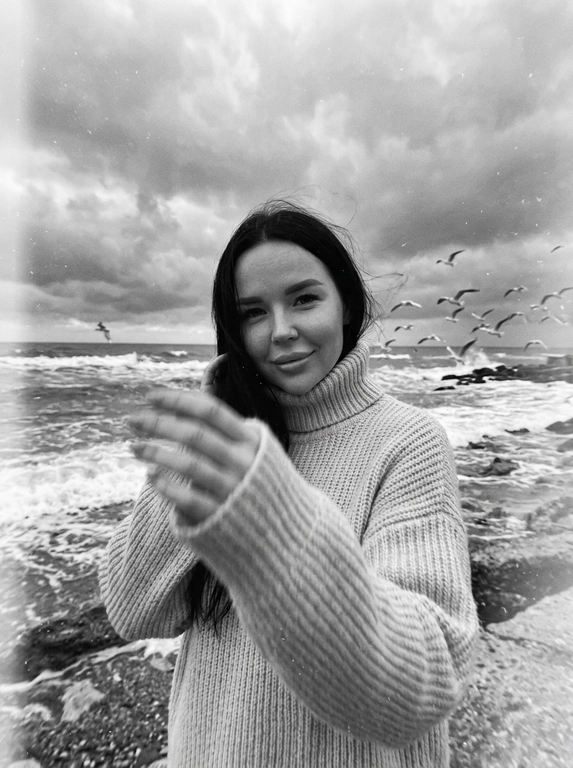 Создай портрет, не меняя черты лица. Черно-белая фотография в стиле ретро: человек стоит на морском побережье, в полупрофиль, смотрит нежно в камеру, легкая улыбка на лице, одет(а) в светлый оверсайз вязаный свитер с высоким воротником. На переднем плане — размытая рука в движении (motion blur), на заднем плане — море с волнами и стая птиц в небе, пасмурное небо. Сильная зернистость пленки, винтажная эстетика, атмосфера свободы, эффект размытия в движении, эстетика.