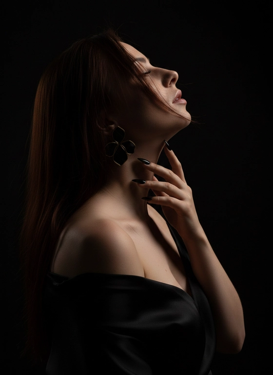 Ultra realistic photo EXACT match to reference pose lighting and composition STRICT identity lock identical face no changes no stylization portrait up to chest woman in profile body slightly turned head thrown back chin lifted up elongated neck pose one hand raised fingers softly touching the neck along the jawline elegant fingers long sharp beige nails large black statement earrings half flower shape with large petals gold outline black satin fabric draped over body open shoulders matte texture hair styled smooth very dense blow dried with a brush loose slightly moving as if from wind one strand falling across the face makeup defined contouring and highlight only subtly visible on rim eyebrows brown laminated eyelashes spiky clustered luchiki lips full extreme low key lighting almost complete darkness face completely swallowed by shadow no visible facial features single very narrow hard rim light strictly from behind and slightly from the side light does not hit the front of the face at all only a thin contour of light visible along silhouette jawline neck shoulders collarbones no fill light no front light no side light no ambient light no bounce light intentionally underexposed image deep black shadows most of the frame nearly black absolute pure black background no details ultra dark cinematic grading crushed blacks minimal warm highlights on skin edges subtle film grain RAW look intimate dramatic almost abstract silhouette aesthetic tanned skin subtle shimmer particles visible on highlighted edges 85mm lens f 1.8 shallow depth of field sharp rim edge rest fades into black, negative prompt front light face lighting visible face illuminated face beauty lighting soft light fill light bounce light ambient light studio lighting bright exposure high key visible details in shadows gray background low contrast flat lighting, лицо нельзя менять, цвет волос, маникюр черный!