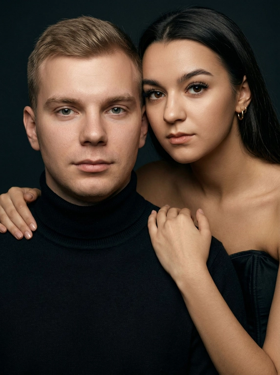 Гиперреалистичный студийный fashion-портрет молодой пары по загруженному референсу, 100% сходство лиц. Черты лица не изменять. Крупный план, кадр по плечи. Мужчина, спокойный уверенный взгляд прямо в камеру, естественное выражение лица, одет в черный трикотажный свитер с высоким воротом. Женщина стоит позади мужчины и нежно обнимает его, ее руки лежат на его плечах. Ее лицо расположено рядом с его лицом, она повернута слегка боком, голова немного наклонена вперед, взгляд направлен прямо в камеру. У женщины выразительные глаза с длинными густыми черными ресницами, естественный clean-glam макияж, натуральные брови, теплый оливковый оттенок кожи, небольшие серьги-кольца. Композиция плотная и интимная, их лица расположены очень близко друг к другу, современный модный портрет пары. Женщина одета в черный корсет. Кожа максимально натуральная: живая текстура кожи, видимые микропоры, subtle skin glow, естественный мягкий блеск кожи, natural skin microtexture, skin translucency, micro skin details, легкие естественные несовершенства кожи, без ретуши и без эффекта пластика. Освещение: мягкий студийный beauty lighting, рассеянный ключевой свет спереди и немного сбоку, мягкие глубокие тени для объема лица, деликатные световые блики на коже. Фон: глубокий темный студийный фон, темно-графитовый или почти черный, создающий контраст с кожей и лицами. Стиль: luxury fashion editorial photography, high fashion portrait, ultra photorealistic, 85mm portrait lens, shallow depth of field, creamy bokeh, cinematic soft lighting, RAW photo, extremely detailed, fine hair strands, natural skin pores, professional photography, photorealism, 8k, high dynamic range.