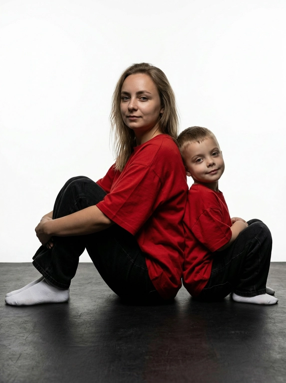 Гиперреалистичная фотография. Качество 8К. Используя портретное сходство с людьми с фотографий, создай фотографию: женщина в красной оверсайз свободной футболке и свободных черных джинсах, белых носках. Ребенок одет в красную оверсайз футболку, черные свободные джинсы и белые носочки. Женщина сидит на полу, согнув колени, смотрит прямо в кадр с еле заметной улыбкой. Сзади женщины в такой же позе сидит ребенок облокотившись спиной к спине женщины, с легкой улыбкой. Темный стиль модного редакционного журнала, глубокие тени и драматическое студийное освещение, белый фон, элегантная и мощная композиция, ультрареалистичный, высокая детализация, художественный портрет.