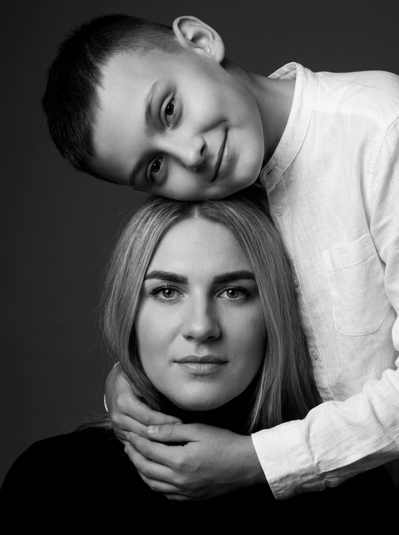 Родитель и ребёнок с внешностью как на приложенных фото, художественная семейная студийная фотосессия. Композиция — вертикальный портрет, крупный план лиц. Родитель внизу смотрит прямо в камеру, лицо фронтально. Ребёнок сверху, голова повернута набок, щека лежит на голове родителя, смотрит в камеру с нежной улыбкой. Рука ребёнка обнимает родителя за шею, видна в кадре, родитель держит руку ребёнка своей рукой.
Одежда: ребёнок в белой льняной рубашке, родитель в чёрной водолазке. 
Фон — тёмно-серый почти чёрный однотонный студийный, создаёт контраст. Освещение — мягкое боковое студийное, создаёт объём и глубину.
Чёрно-белая художественная фотография с выразительными контрастами. Атмосфера нежная, интимная, спокойная.