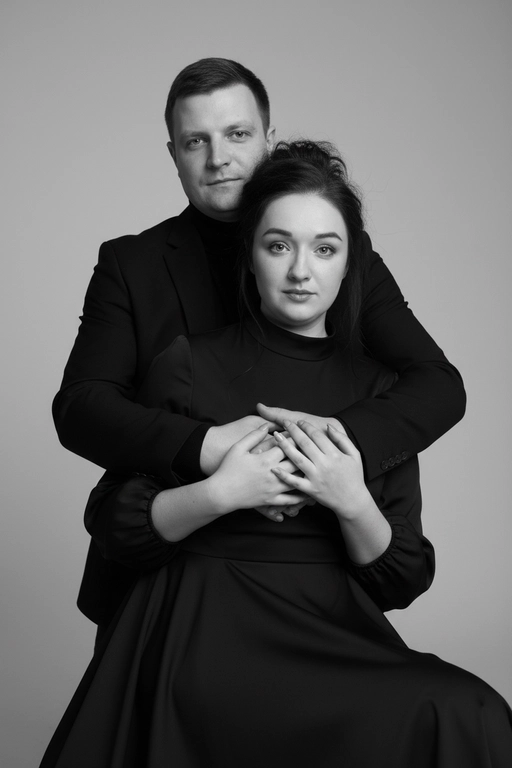 Черно-белый портрет мужчины и женщины в стиле фэшн фотосессии в фотостудии. Не изменяйте лица. Фон нейтральный, однотонный, светло-серый. Атмосфера интимная и спокойная. На переднем плане женщина. Вся композиция выполнена в приглушенной монохромной гамме со студийным освещением, которое подчеркивает текстуру кожи и одежды. Её руки лежат на груди мужчины. Мужчина обнимает женщину сзади, положив свою руку поверх её руки. Она одета в вечернее платье, у нее красивая прическа и макияж. Мужчина одет в  классическом черном пиджаке и черной водолазке.