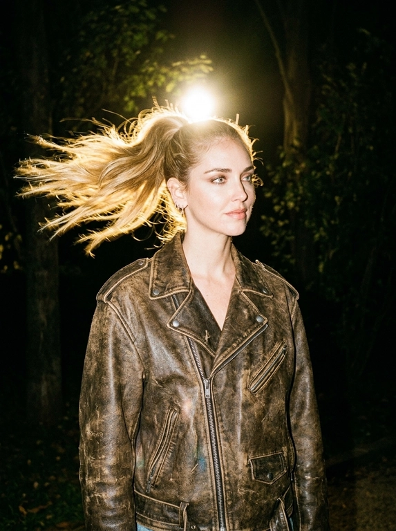 Ultra-realistic nighttime candid fashion photograph, recreate the reference composition precisely. Medium portrait outdoors at night. The woman stands slightly turned to the side, torso leaning subtly forward, head slightly tilted, eyes looking sideways with a calm confident expression.
She wears an oversized vintage BROWN LEATHER BIKER JACKET with wide shoulders, metal zipper details and heavily distressed worn leather: faded patches, rubbed edges, natural creases, scratches and authentic aged patina. Fitted black top underneath.
Nighttime outdoor setting with trees disappearing into darkness. An extremely bright warm light directly behind her head creates a powerful golden halo. Strong backlight creates warm golden-orange rim lighting around her silhouette. Direct camera flash softly illuminates her face and brown jacket from the front.
Deep black surroundings, intense golden backlight, warm lens flare, slight overexposure around the light source. Raw late-night 2000s flash photography, slightly imperfect sharpness, digital noise, warm vintage color grading, candid spontaneous atmosphere, realistic photography, no AI look.

Strong wind sweeps the hair dramatically sideways, spreading it into a flowing fan behind her. Individual strands are naturally separated and slightly tousled. Golden backlight shines through the moving strands, with subtle natural motion blur only in the hair.

Причёска: Hair gathered into a high ponytail with soft volume at the crown.