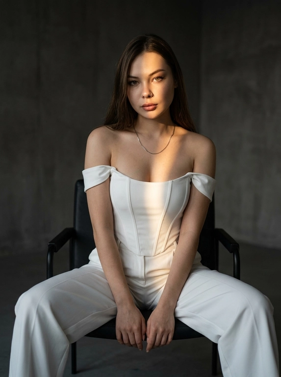 Фотореалистичная fashion-фотография женщины, внешность и черты лица максимально точно сохранить по загруженному референсу, возраст не менять. Женщина сидит в расслабленной уверенной позе на современном чёрном кресле, ноги широко расставлены, корпус слегка наклонён вперёд, предплечья лежат ближе к коленям, кисти рук свободно соединены перед собой. Минималистичная тёмно-серая студия, вокруг почти нет декора.

Главный художественный акцент — узкая полоса тёплого закатного солнечного света, проходящая диагонально через лицо, плечо и верхнюю часть груди. Одна часть лица ярко подсвечена золотистым солнцем, другая остаётся в глубокой мягкой тени. Очень выразительный светотеневой рисунок, cinematic chiaroscuro, драматичный контраст, глубокие тени, тёплые блики на коже. Спокойный уверенный взгляд прямо в камеру, слегка серьёзное выражение лица. Реалистичная кожа с естественной текстурой и порами, без пластиковой ретуши. Editorial fashion photography, luxury minimalism, cinematic mood, 50mm lens, f/2.0, ISO 100, высокая детализация, натуральные пропорции тела.

Какая будет одежда?: На женщине белый облегающий корсетный топ без бретелей с чёткими вертикальными швами и свободные белые брюки с высокой посадкой. Образ выглядит чисто, минималистично и дорого, с акцентом на графичный силуэт и контраст света и тени.
