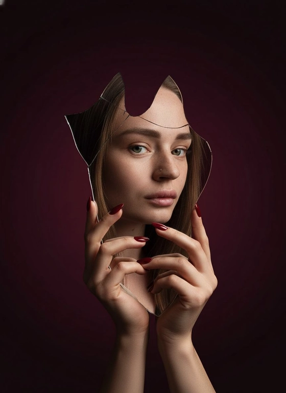 Создай фото для человека на предоставленном фото. Не меняй черты лица. Осколок разбитого зеркала держат изящные руки с длинными красными маникюром, отражая лицо человека. Выражение её лица серьёзное и выразительное. Фон — плавный градиент тёмно-красного цвета для эффектного контраста. Кинематографическое освещение с глубокими тенями, резкий фокус на глазах, сюрреалистичный и художественный стиль модной фотографии.