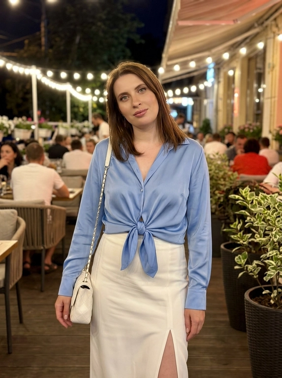 Фотореалистичное фото, снятое на iPhone, в эстетике elegant night outfit, feminine luxury vibe, Pinterest style. Кадр по бёдра или чуть ниже, модель находится на фоне стильного вечернего кафе или летней террасы. Фото выглядит естественно, как живой городской кадр без студийной постановки.

Модель с вечерним glam-макияжем: кожа с натуральной текстурой, длинные ресницы, подчёркнутые глаза, мягкий контуринг и розовые глянцевые губы. Выражение лица спокойное, мягкое, отстранённое и уверенное.

На модели через плечо белая стёганая сумка на цепочке. Образ элегантный, дорогой, вечерний и женственный.

Модель стоит в расслабленной fashion-позе: корпус слегка изогнут, одна рука заведена назад или лежит у талии, другая естественно держит сумку на плече. Голова немного наклонена в сторону, взгляд мягкий и уверенный. Поза плавная, женственная, с акцентом на талию, разрез юбки, сумку и силуэт.

На заднем плане вечерняя летняя терраса или вход в красивое кафе: мягкий тёплый свет лампочек сверху, цветы в кашпо, светлый декор, стеклянные панели, уютная ночная городская атмосфера. Фон слегка размыт, но читается как стильное модное заведение.

Освещение мягкое ночное, смешанное: тёплые лампы сверху и лёгкая холодноватая подсветка заведения на фоне. Свет не выглядит как студийный свет.

Цветокор cinematic: глубокие мягкие тени, сочетание голубовато-белых и тёпло-золотистых оттенков, лёгкий контраст, натуральные цвета, без чрезмерного HDR и без глянцевой рекламной обработки. Естественная текстура кожи, soft shadows, real life shot, iPhone photo, casual luxury lifestyle, no studio lighting, no CGI, no plastic skin, no over-retouching.

Какая будет одежда?: Нежно-голубая атласная блуза с длинными рукавами и глубоким V-образным вырезом, завязанная узлом спереди. Длинная белая юбка с высокой посадкой и глубоким разрезом на бедре.

Какая будет прическа?: Не менять мою прическу
