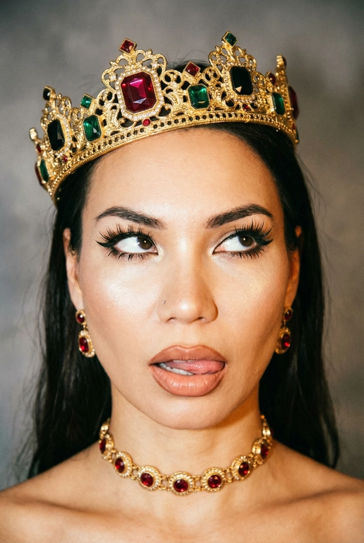 Создай фото -детальное описание художественного портрета девушки, выполненного в популярной современной эстетике, сочетающей элементы «е-qirl» и стиля «rebellious royalty» (бунтарская королевская особа).
:Центральный образ и внешность:
• Лицо и поза: Девушка позирует в дерзкой и провокационной манере.
Её глаза закатаны (или направлены сильно в сторону и вверх), а кончик языка слегка высунут. Это создает образ, сочетающий в себе игривость, бунт и некоторую отрешенность.
Элегантный макияж глаза с длинными острыми черными стрелками, длинные густые ресницы эффект лучики, нижние ресницы накрашены, хайлайтер во внутренних уголках и скулах.
Пухлые полностью прокрашенные телесной матовые губы слегка приоткрыты легкий блеск и светло-коричневый карандаш по краям. персиково-розового оттенка.
Не меняй мою прическу. 
Корона: На голове массивная высокая корона в винтажном стиле. Она украшена сложным филигранным узором и крупными драгоценными камнями: по центру расположен большой квадратный красный камень (рубин), по бокам - камни зеленого (изумруд) и черного цветов. Корона сидит немного небрежно, подчеркивая неформальность образа.
Ожерелья:
На шее надет чокер, выполненный в одном стиле с короной - цепочка с круглыми красными камнями в оправе. В ушах серьги по такому же типо как корона •
Фон: Нейтральный, дымчато-серый или слегка туманный. Он лишен деталей, что позволяет полностью сфокусировать внимание на ярких аксессуарах и лице модели.
Освещение и обработка: Свет мягкий, рассеянный. Фотография имеет характерную обработку с эффектом «зернистости» (пленочное зерно), что придает ей винтажный и художественный вид.
Цветовая гамма теплая, с акцентом на золото и красный цвет.
Общее впечатление:
Снимок транслирует идею современной интерпретации власти и красоты. Это противопоставление классического символа величия (короны) и современного неформального поведения.
Образ выглядит стильно и кинематографично и запоминающе, сильная вспышка. Корона цвета золота.