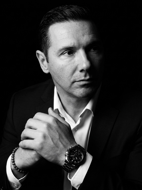 A hyper-realistic 8K cinematic portrait of a uploaded person,
captured in a dramatic Chiaroscuro style. The subject is seated at a
three-quarter angle, leaning slightly forward with a relaxed yet
commanding posture. His face is turned away from the lens, with
one side of his face clearly illuminated while the other is
completely lost in deep, velvety black shadows. His expression is
contemplative, not looking at the camera. The pose is precise: his
hands are clasped together near his chest; one hand is held
straight with fingers slightly tilted, while the other hand is
positioned so only the fingers are visible, resting over the first. He
wears a luxury black chronograph watch with a detailed metal link
bracelet, and a beaded bracelet. His attire is a sharp
black suit jacket over a white dress shirt with the top buttons
open, showing refined fabric textures. The background is a solid,
seamless black. The lighting is sharp and directional, highlighting
the intricate details of his hair,
the moisture in his eyes, and the glint off the watch face.
Cinematic black-and-white color grade with rich contrast and
smooth, natural skin transitions. Shot on 85mm lens, f/1.8, high-
resolution commercial photography