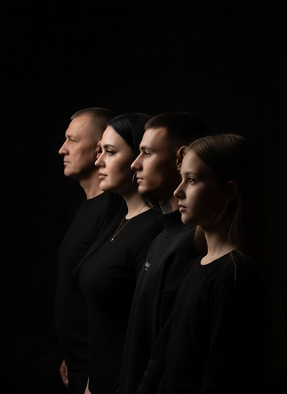 Профессиональный студийный портрет с низким контрастным светом. Головы расположены строго в одну линию, каскадом. боковой профиль, взгляд влево. У всех видны только головы и немного плечи. Черный фон. Черная одежда у всех. Люди занимают весь кадр. 4 человека. Порядок слева на право: отец-> мать-> сын-> дочь.