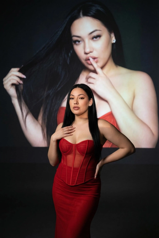 Создайте фотографию женщины в элегантной позе. На ней — слегка прозрачный облегающий красный корсет и длинное облегающее красное платье. Одной рукой она касается области груди, а другой — мягко опирается на бедро, что создаёт динамичную позу. Она смотрит с уверенностью и чувственностью. На ней простые серьги.
Эта женщина на переднем плане занимает меньшую часть высоты изображения. На ней — слегка прозрачный облегающий красный корсет и длинное облегающее красное платье.
Фон тёмный и минималистичный, с мягкой теневой проекцией той же женщины (в другой позе или в виде абстрактной фигуры) в верхней части изображения. Проекция — естественное цветное изображение женщины, которая представлена значительно крупнее и занимает большую часть верхней части фона. Она выглядит соблазнительно: одной рукой держит волосы, а палец другой руки прижала к губам, смотря при этом соблазнительным взглядом.
Не изменяй черты лица. Длинные прямые  черные волосы