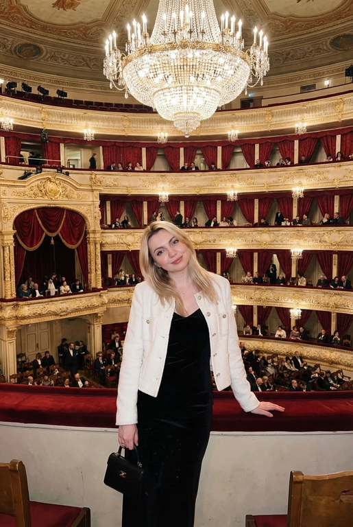 Я стою в ложе Большого театра, на рядов лож и большой хрустальной люстры. В ложах сидят люди, висят красные бархатные занавески. Камера на уровне глаз.  Съёмка выглядит как фото, сделанное на iPhone: естественный свет, лёгкое зерно, минимальная обработка, живой момент без позирования. Взгляд в камеру, легкая улыбка. Голова немного наклонена вбок. Волосы сияют, макияж естественный, губы глянцевые. Одна рука лежит на бортике ложа, во второй - маленькая черная сумочка. Приглушенный свет. На мне чёрное бархатное вечернее платье без рукавов по фигуре и белый жакет в стиле Channel. 