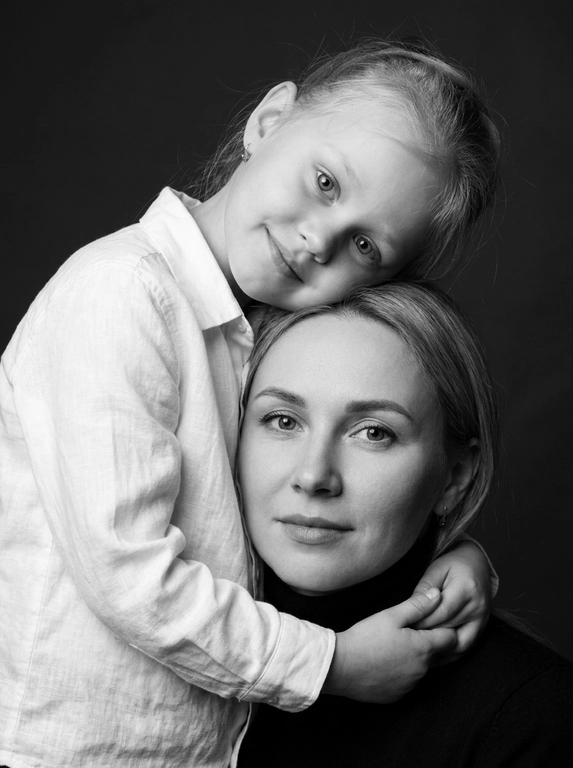 Родитель и ребёнок с внешностью как на приложенных фото, художественная семейная студийная фотосессия. Композиция — вертикальный портрет, крупный план лиц. Родитель внизу смотрит прямо в камеру, лицо фронтально. Ребёнок сверху, голова повернута набок, щека лежит на голове родителя, смотрит в камеру с нежной улыбкой. Рука ребёнка обнимает родителя за шею, видна в кадре, родитель держит руку ребёнка своей рукой.
Одежда: ребёнок в белой льняной рубашке, родитель в чёрной водолазке. 
Фон — тёмно-серый почти чёрный однотонный студийный, создаёт контраст. Освещение — мягкое боковое студийное, создаёт объём и глубину.
Чёрно-белая художественная фотография с выразительными контрастами. Атмосфера нежная, интимная, спокойная.