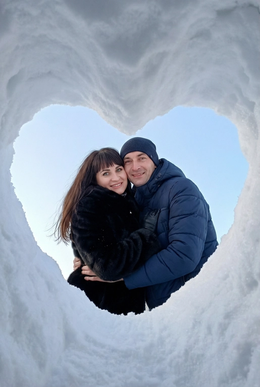Фотография этой пары, снятая снизу вверх через отверстие в снегу в форме сердца. Камера находится в снежной нише, края кадра — плотный белый снег, образующий чёткое сердце. В центре кадра двое человек из референса, обнимающие друг к друга, смотрят прямо в камеру. Люди одеты по-зимнему.   Их одежда стильная и современная. Они улыбаются, лица хорошо видно. 
Фон — чистое светло-голубое зимнее небо без облаков. Естественный дневной свет, мягкие холодные оттенки, высокая детализация снега и ткани одежды. Атмосфера — уют, любовь, спонтанный зимний момент. Реалистичная фотография, без искажений лиц, фокус на людях, эффект живого кадра, как с экшн-камеры или смартфона, вертикальный формат. Женщина одета в черную короткую шубу , волосы распушены ушей не видно , чёлка. Глаза голубые с зеленцой, мужчина в темно синий пуховик и темную шапку.