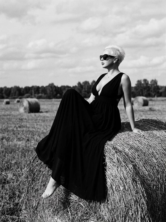 Чёрно-белая high-fashion editorial фотография в сельской местности, женщина с внешностью как на референсе, черты лица не изменять, композиция — более общий план (wide shot), камера на среднем расстоянии, фигура полностью в кадре с окружающим пространством, она сидит на большом тюке сена в поле, корпус слегка развернут, одна нога свободно опущена вниз, вторая согнута, создавая расслабленную, но выразительную позу, руки естественно лежат на колене и на поверхности тюка, взгляд направлен в сторону, профиль, выражение спокойное и уверенное, одежда: чёрный топ с V-образным вырезом и объёмные светлые широкие джинсы с потёртостями и рваными элементами, аксессуары: крупные солнцезащитные очки, минималистичные украшения, фон: открытое поле с несколькими тюками сена на разной глубине, создающими перспективу, линия горизонта ниже центра кадра, чтобы подчеркнуть небо, освещение: жёсткий естественный дневной свет с мягкими облаками, формирует выразительные тени и контраст, стиль: кинематографический, плёночное зерно, высокая детализация, реалистичная текстура кожи и ткани, редакционный fashion стиль, акцент на атмосфере и пространстве кадра


Какая будет прическа?: НЕ изменять мою прическу

Доп. характеристики: Девушка в красивом длинном чёрном платье с декольте
