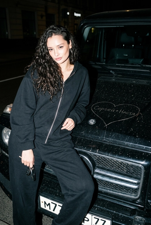 Строго сохранить лицо с прикреплённой референс-фотографии, на 100% без искажений. Человек одет полностью в чёрный, монохромный оверсайз спортивный костюм: свободная худи на молнии и широкие расслабленные штаны, без логотипов, текста и брендинга. Акцент на реальной текстуре кожи и детализированной текстуре ткани.
Сырой кинематографичный ночной кадр. Человек опирается на капот автомобиля, средний план, лёгкий верхний ракурс. В одной руке игриво держит ключи от машины. Номерной знак виден: m777yp 77.
Строго по центру капота, покрывая всю поверхность капота, глубоко и грубо процарапаны (не мелом) царапины: большое сердце с текстом «» внутри.
Жёсткая прямая вспышка, сильные тени, высокий контраст, видимая пыль и микро-царапины на краске. Тёмный размытый городской фон.
Объектив 35mm, f/2.8, грубая уличная эстетика.

Надпись на капоте: Сергей+Настя

Марка автомобиля: Опирается на чёрный Mercedes-Benz G-Class