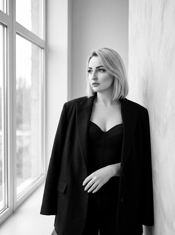 Женщина слегка наклонилась к стене в углу комнаты. Сохрани лицо и черты лица с оригинального фото. На ней надет большой чёрный пиджак, накинутый на плечи, и чёрный лаконичный топ-корсет с V-образным вырезом. Женщина стоит, смотря в сторону окна вдаль, выражение лица спокойное и уверенно задумчивое. Она слегка опирается на стену, положив руку на пиджак. Комната светлая, с большими окнами слева, пропускающими естественный свет. Общая атмосфера спокойная и стильная, подчёркивающая элегантность и минимализм. Положение женщины в кадре — справа от центра. Освещение мягкое, естественное, создающее ощущение минимализма и простоты. Фотография выполнена в чёрно-белом стиле, подчёркивающем контрасты и текстуры. Атмосфера спокойная, элегантная и немного загадочная. Максимально близкий ультрареалистичный 8K портрет.
