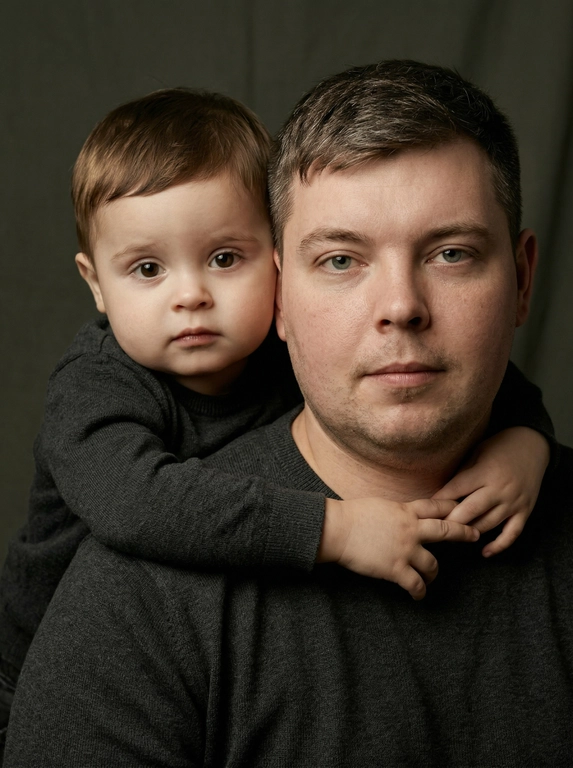 Создай портретный студийный кадр из фотосессии отца и сына. Сын обнимает отца сзади, лежа подбородком на его плече, руки мягко сомкнуты на груди мужчины. Оба смотрят прямо в камеру. Естественные спокойные эмоции, лёгкая серьёзность во взгляде.
Одежда — однотонные чёрные лонгсливы или свитера без принтов. Тёмный нейтральный фон (глубокий серо-оливковый или графитовый). Мягкий направленный студийный свет с лёгкой теневой глубиной, акцент на лицах, тёплая естественная цветокоррекция, высокая детализация кожи, натуральная текстура без излишней ретуши.
Кадр — крупный или средний план, плотная композиция, лица расположены близко друг к другу. Атмосфера — тёплая, камерная, эмоциональная, ощущение близости и доверия. Профессиональная съёмка, 85mm lens, shallow depth of field, cinematic portrait, ultra realistic.