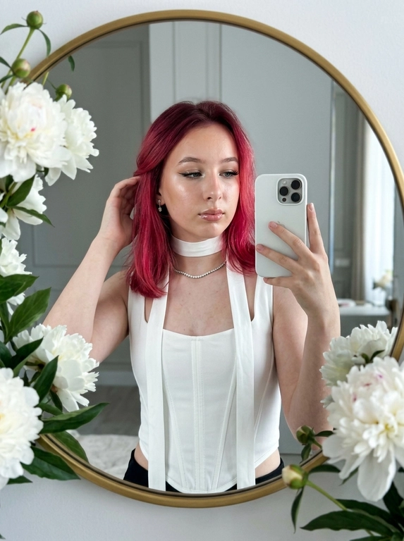 Максимально реалистичное селфи в зеркале в эстетике luxury soft glam. Модель стоит перед круглым зеркалом с изящной рамой, украшенной белыми живыми пионами. Цветы обрамляют зеркало нежно и дорого, создавая ощущение утончённого, премиального кадра. Композиция чистая и визуально собранная: модель находится в центре отражения, кадр выглядит эстетично, дорого и современно.

Поза естественная и уверенная: одной рукой модель держит современный смартфон, второй рукой слегка касается волос, создавая живой и непринуждённый жест. Селфи выглядит не постановочным, а как красивый спонтанный кадр из дорогой life-style съёмки.

Макияж — выразительный soft glam: чёткие чёрные стрелки, длинные пышные ресницы с лисьим эффектом, внутренние уголки глаз подсвечены сияющими тенями. Кожа ровная, ухоженная, с мягким влажным сиянием. Губы подчёркнуты карандашом и покрыты полупрозрачным глянцевым блеском, создающим аккуратный объём.

На модели ее образ выглядит женственно, современно и дорого. 

Смартфон современный, в минималистичном белом матовом чехле. Если нужен более дорогой вайб, без лишнего декора и ярких деталей.

Освещение холодное, мягкое и чистое, как в дорогой beauty-съёмке или premium dressing room. Свет красиво подчеркивает сияние кожи, текстуру ткани, свежесть пионов и блеск украшения. Атмосфера — роскошная, женственная, ухоженная, без дешёвого глянца.

Фотореализм максимальный: высокая детализация кожи, ткани, цветов, аксессуаров и отражения в зеркале. Лёгкий цифровой шум, мягкая резкость, деликатная зернистость, естественная глубина кадра, высокая контрастность, эффект живого фото, без CGI и без искусственного AI-эффекта.

Какая будет прическ?: Не менять мою прическу

Какой будет образ на фото? : Белый топ-корсет с открытыми плечами. Его главная особенность — длинная лента (или шарф), выполненная из той же ткани, что и топ. Лента плотно охватывает шею, создавая эффект шарфа, прямые и свободные концы ленты спускаются вниз они проходят вдоль передней части топа, свободно свисая.  Образ элегантный.  Акцент на линии шеи и вертикальных линиях торса.