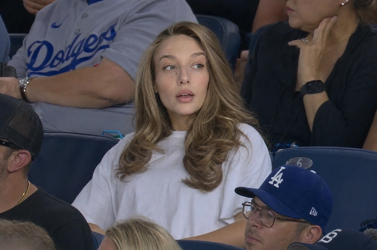 Person sitting in the crowd at a professional baseball game (or it could be any other match that I will write below), filmed from far away by a live broadcast camera. 
Seated in blue stadium chairs, slightly leaned back, looking off to the side with a surprised caught-on-camera expression, lips slightly parted.

Какая будет одежда? : Не менять мою одежду