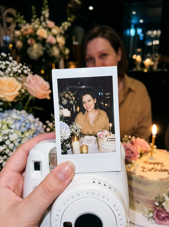 Edit the uploaded reference image into a photorealistic, luxury
birthday party scene.
Preserve the person’s identity exactly. Keep their real facial
structure, skin tone, age presentation, hair type, body type, natural
proportions, expression style, and distinctive features. The final
image should still clearly look like the same person from the
reference photo.
Create the image in the style of a dark, intimate iPhone UGC birthday
snapshot taken in a dim room with direct front flash. Use a dynamic
handheld camera angle, slightly tilted or off-center, as if the photo
was taken spontaneously at the party. The perspective should feel
natural, candid, and immersive, with the white instant camera with
polaroid angled and in focus of the generated image. Avoid a stiff,
perfectly centered studio composition.
The mood should feel candid, and special, like a real birthday
moment captured casually at night. Use flash-lit foreground details,
darker background shadows, warm candle glow, subtle phone-
camera grain, slight handheld imperfection, and shallow depth of
field.
The main focus must be a freshly printed instant polaroid photo
coming out of a clean white instant camera in the foreground. The
white camera should be large in the lower foreground, held close to
the lens. The polaroid should be sharp, visually dominant, and
positioned with a natural slight angle for a more dynamic
composition. The composition should feel similar to a casual iPhone
photo where the polaroid and camera are the main subject, while the
actual person behind it is only subtly visible, and not the main focus,
face not visible.
A natural, well-groomed hand matching the person’s skin tone and
overall styling holds the white instant camera. The hand should look
realistic and relaxed, with simple polished nails only if suitable for
the person.
Inside the polaroid, show the same person from the reference image
posing elegantly at their birthday celebration. They should look
confident, refined, and warmly expressive. Transform their outfit into
a polished birthday look that suits their appearance and gender
presentation. For a woman, create an elegant dress or refined
celebratory outfit. For a man, create a fashionable formal or semi-
formal outfit suitable for a luxury birthday setting. The outfit color should
harmonize with or be inspired by the outfit color in the original
reference photo.
Behind the polaroid, show only a subtle blurred glimpse of the same
person in the background, wearing the same transformed outfit. The
background person should be softly out of focus, partly hidden by
the polaroid camera.
Include tasteful birthday décor around the scene. Use elegant
flowers that harmonize with the subject’s outfit and the overall color
palette of the reference photo. The flowers may be roses, orchids,
hydrangeas, baby’s breath, peonies, lilies, greenery, or other refined
florals. Choose colors that match the person’s transformed outfit
and create a cohesive luxury birthday atmosphere.
Place a birthday cake to the side or slightly in the background. The
cake should be visible but secondary to the polaroid. The cake color,
icing, floral decorations, and candle color should harmonize with the
person’s outfit and the overall palette. Add one lit candle. The cake
must clearly say exactly: “Happy Birthday” in neat, readable icing.
Use realistic direct flash highlights, intimate dark-room lighting, warm
candlelight, soft party lights, shallow depth of field, realistic skin
texture, natural facial proportions, elegant styling, and a cohesive
color palette based on the reference image.
**Strict constraints:** no identity change, no altered facial structure,
no mismatched person between the polaroid and background, no
extra people, no rings, no bracelets, no hand jewelry, no distorted
hands, no extra fingers, no duplicated faces, no warped camera, no
unreadable cake writing, no misspelled cake text, no extra text, no
overly bright studio lighting, no flat commercial lighting, no plastic
skin, no stiff straight-on composition