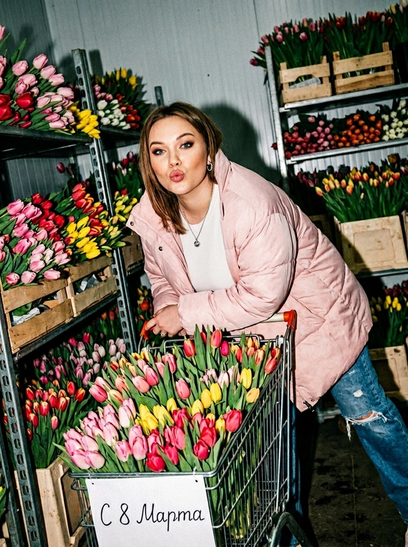 Создай фото, не меняя черты лица. Женщина толкает тележку, переполненную цветов. На тележке табличка «С 8 Марта». На ней большой нежно-розовый пуховик, расстёгнутый, голубые джинсы с потертостями, топ. Безупречный макияж. Она наклоняется вперёд над ручкой тележки с игривым выражением лица, словно посылая поцелуй в сторону камеры. За ней — полки, заставленные теми же цветами. Непринуждённый, естественный, спонтанный стиль фотографии. Съёмка с резкой прямой вспышкой — сильные тени, пересвеченные участки, неравномерная экспозиция. Наклонная композиция: объекты не по центру, обрезанные конечности, неидеальное выравнивание. Размытие в движении, неидеальная фокусировка, съёмка в середине движения или в середине выражения эмоций. Шум и зернистость — без ретуши или сглаживания. Небрежная, интимная, реалистичная, документальная атмосфера.


Какие будут цветы?: Тележка полная тюльпанов. Полки заставленные тюльпанами.