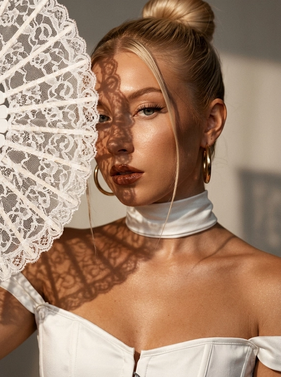 Ultra realistic beauty editorial portrait. Крупный портрет модели: лицо, шея и плечи, лёгкий наклон камеры. Белый кружевной веер частично закрывает лицо, создавая чёткие lace shadows на щеке, глазе, шее и ключице.
Белый структурный корсет с открытыми плечами и плотный satin neck wrap, плотно облегающий шею.  Макияж: sharp eyeliner, длинные ресницы, glossy brown lips, сияющая бронзовая кожа с натуральной текстурой.
Тёплый направленный свет, глубокие тени, золотистый glow, крупные золотые серьги, cinematic contrast, subtle film grain, Vogue aesthetic, RAW realism, ultra detailed 8K, no plastic skin.

Какая будет прическа?: Волосы — sleek high bun с тонкой прядью у лица.