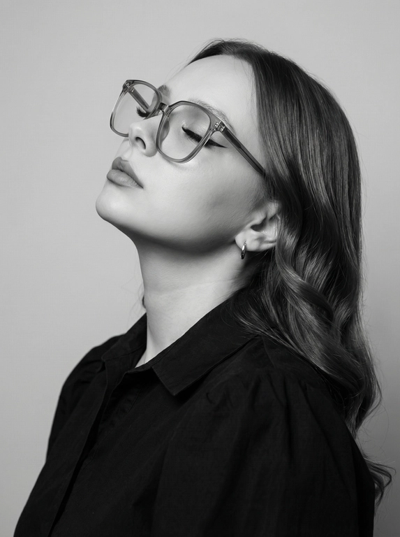 Чёрно-белый минималистичный фотореалистичный портрет, студийный мягкий свет спереди‑сверху, нейтральная светлая однотонная стена как фон. Девушка в профиль: голова запрокинута назад, подбородок поднят, глаза закрыты — расслабленная fashion-поза с акцентом на линии шеи и контуры лица. Обязательно точно сохранить черты лица с референс-фото (без искажений); крупные квадратные очки в массивной полу-прозрачной оправе с естественными бликами. Детали лица: длинные ресницы и чёткие стрелки, выразительные губы, естественная текстура кожи, аккуратный ретуш. Одежда: тёмный объёмный верх с выразительным воротником, маленькая серьга-кольцо. Стиль: высокая графичность и контраст, чистые линии, лёгкая драматичность, максимальная детализация и фотореализм, аккуратные светотеневые переходы, подчёркнутые контуры.

Какая будет прическа?: Волосы уложены в гладкие локоны
