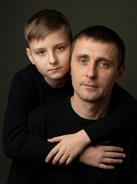 Создай портретный студийный кадр из фотосессии отца и сына. Сын обнимает отца сзади, лежа подбородком на его плече, руки мягко сомкнуты на груди мужчины. Оба смотрят прямо в камеру. Естественные спокойные эмоции, лёгкая серьёзность во взгляде.
Одежда — однотонные чёрные лонгсливы или свитера без принтов. Тёмный нейтральный фон (глубокий серо-оливковый или графитовый). Мягкий направленный студийный свет с лёгкой теневой глубиной, акцент на лицах, тёплая естественная цветокоррекция, высокая детализация кожи, натуральная текстура без излишней ретуши.
Кадр — крупный или средний план, плотная композиция, лица расположены близко друг к другу. Атмосфера — тёплая, камерная, эмоциональная, ощущение близости и доверия. Профессиональная съёмка, 85mm lens, shallow depth of field, cinematic portrait, ultra realistic.