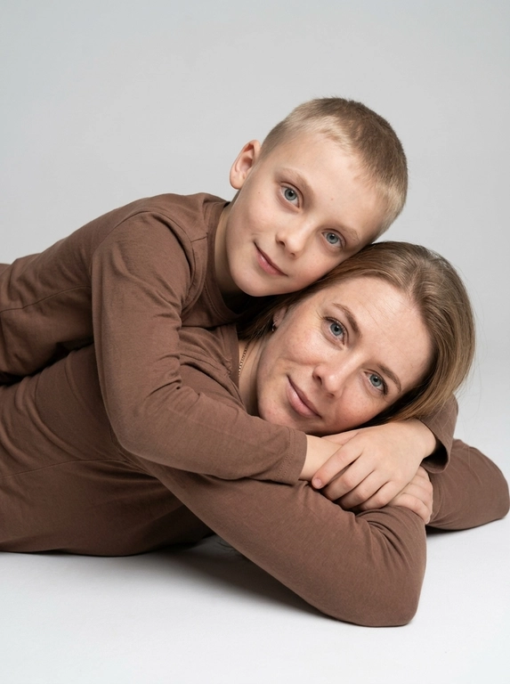 Создай студийный портрет родителя и ребёнка, максимально точно сохранив черты лица и выражения с референсных изображений. Не изменяй и не искажай лица, воспроизведи их с фотографической точностью.

Композиция:

Крупный план, ракурс на уровне глаз.

Родитель и ребёнок лежат на животе, расположены боком к камере.

Ребёнок лежит на спине родителя и мягко обнимает его за плечи.

Оба смотрят прямо в камеру, создавая тёплый и близкий зрительный контакт.

На родителе и ребёнке одинаковые коричневые лонгсливы без принтов и декора.

Естественный внешний вид, ровный тон кожи.

Мягкое студийное освещение, спокойная семейная атмосфера, натуральная текстура кожи, высокая детализация, максимальный фотореализм. Без ламп в кадре и прочего декора.

Формат 9:16.

Качество 8K.