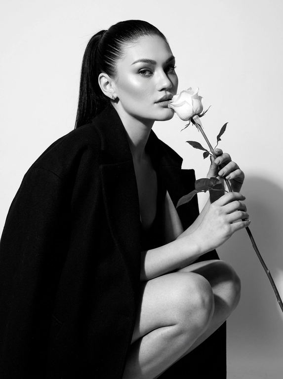 Высоко модная черно-белая редакционная фотография женщины в эффектной позе. Она грациозно присела на корточки, накинув на плечи объемное структурированное черное пальто, оставляя ноги открытыми.
Держит в руке белую розу с длинным стеблем и листьями, поднося к губам бутон. Изящный, минималистичный стиль. Сильное направленное студийное освещение, создающее глубокие тени и высокий контраст. Изысканный макияж с четкой подводкой для глаз и блеском на губах. Чистый фон, эстетика роскошного журнала, стиль Vogue, сверхчеткая, высококонтрастная монохромная фотография. Не изменяйте и не модифицируйте структуру лица, тон кожи или пропорции тела модели.
Только освещение, фон и застывшие текстуры должны быть переосмыслены в контексте задания. Сверхчеткое изображение, высокий динамический диапазон, студийная четкость, реалистичная текстура кожи, естественные поры, едва заметная зернистость пленки, редакционная цветокоррекция, съемка для печати.

Какая будет прическа?: Волосы собраны в гладкий хвост