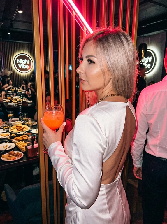 Ресторан. На этой фотографии запечатлена женщина в обстановке ночного ресторана, снимок сделан со спины в пол-оборота, лицо в профиль, что создает динамичный и выразительный образ. На женщине белое атласное платье с открытой спиной, этот наряд подчеркивает фигуру и соответствует вечернему дресс-коду. В правой руке она держит высокий стакан с напитком оранжевого цвета (возможно, коктейль), на шее можно заметить золотое украшение. Макияж — длинные ресницы с эффектом «лучики», губы накрашены блеском. Маникюр короткий квадрат жемчужного цвета. Женщина стоит у барной стойки или декоративной перегородки с вертикальными золотистыми рейками, слева на фоне видны накрытые столы с закусками и тарелками, а также круглая вывеска с надписью Night Vibe (вероятно, название заведения). В помещении приглушенный клубный свет, на фигуру ложится яркий розово-красный блик от неонового освещения или прожектора, что добавляет кадру сочности и атмосферности ночной жизни. Справа в кадр частично попал другой человек в белой рубашке, что подчеркивает многолюдность и социальный характер обстановки. Это классический пример фотографии для социальных сетей, передающий эстетику ночного города и отдыха, эстетика Instagram, черты лица полностью сохранены как на исходном фото.