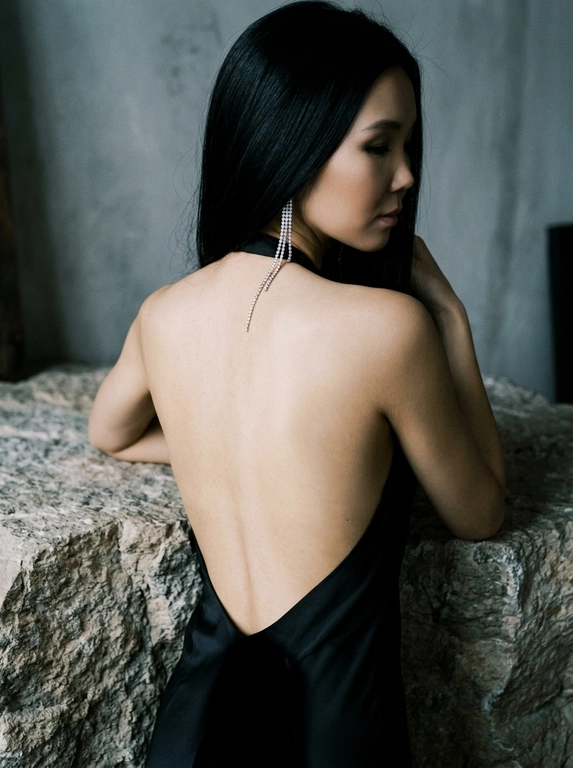 Создай изображение женщины с фотографии, не изменяя лицо, черты, внешность, возраст и индивидуальность. Она в элегантном черном вечернем платье с открытой спиной, ткань подчеркивает тонкую кожу с естественным свечением, складки и блеск шелка придают глубину. Опирается на необработанную каменную стойку с шероховатой фактурой, контрастирующей с платьем и кожей. Приглушенный свет лаунжа создает мягкие тени, подчеркивая изгибы спины и переходы света на платье и камне. Длинная сверкающая диамантовая серьга спускается по спине, ловя свет и создавая мерцание. фото в высоком разрешении с зернистой пленочной текстурой и высоким контрастом, в стиле классического нуара и модной редакционной фотографии, с холодным глубоким визуальным стилем свет усиливают глубину и светотеневой рисунок. Строго сохранить лицо, идентичность и естественную внешность человека с оригинала. Фото в цвете.

Какая будет прическа?: Не менять мою прическу