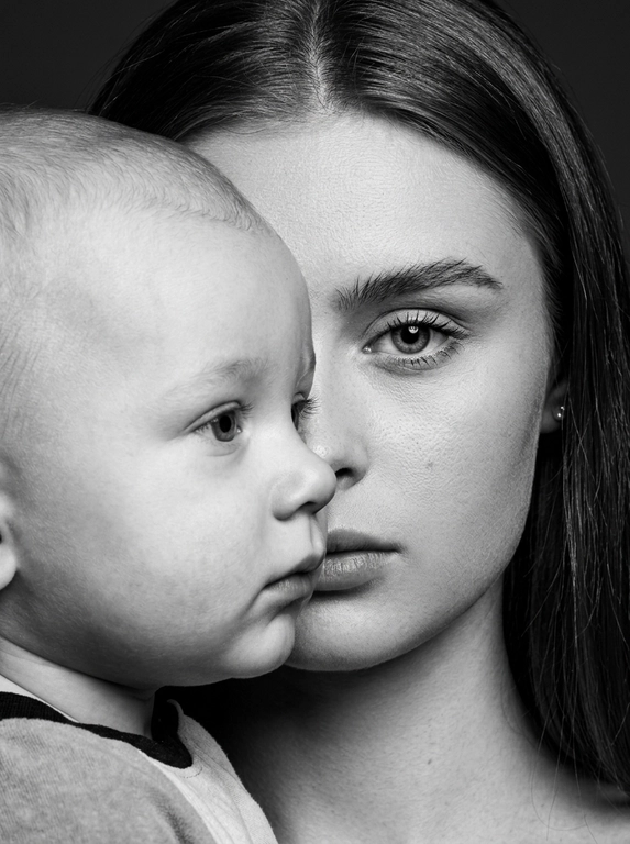 Чёрно-белый фотореалистичный студийный beauty-fashion портрет родителя и ребёнка с представленных фото. Крупный плотный кадр по плечи, без свободного пространства. Ребёнок находится на переднем плане, ближе к камере, в профиль, расположен сбоку и частично перекрывает лицо родителя. Лицо ребёнка закрывает примерно половину лица родителя, создавая сильный эффект визуального перекрытия. Родитель находится позади ребёнка, строго по центру кадра; у родителя виден только один глаз, направленный прямо в камеру, вторая часть лица скрыта лицом ребёнка. Лица расположены максимально близко, почти соприкасаются, но без явного физического действия. Ребёнок смотрит в сторону, выражение спокойное, сдержанное и закрытое. Родитель смотрит прямо в камеру одним открытым глазом, взгляд холодный, спокойный и пронизывающий. Атмосфера high-fashion editorial: минимальная эмоциональность, люксовая отстранённость, скрытое напряжение и холодная близость. Фон тёмный, чистый, студийный, без деталей. Жёсткое студийное освещение, высокий контраст, глубокие тени, выразительная светотень на лицах, натуральная текстура кожи, sharp focus, dramatic black and white photography, editorial beauty portrait, без улыбок, без лишних предметов, без текста и логотипов.