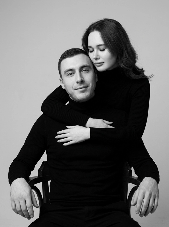 Черно-белая студийная портретная фотография в стиле минимализма. На снимке изображены мужчина и женщина, позирующие на нейтральном светлом фоне. Мужчина сидит на стуле лицом к камере, его руки расслабленно лежат на подлокотниках, взгляд направлен прямо перед собой с мягкой полуулыбкой. Женщина стоит сзади, нежно обнимая мужчину за грудь руками, а ее голова аккуратно прислонена щекой к макушке мужчины с закрытыми глазами. Оба одеты в одинаковые черные водолазки с длинными рукавами. Атмосфера кадра интимная, теплая, спокойная и эмоционально близкая. Цветовая гамма монохромная, выдержанная в контрастных черно-серых тонах с мягким студийным освещением, подчеркивающим текстуру одежды и контуры лиц.