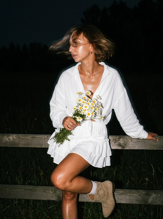 Cinematic candid night shot. iPhone RAW, direct flash, film grain aesthetic.

Girl standing leaning her back and elbows on a wooden fence in a field, distant trees behind her. She suddenly turned her head sharply toward the camera — motion blur from the sharp head turn: hair blur sweeping across the frame, slight blur on face edges, face still recognizable, expression caught mid-movement, alive and unexpected.

hair blown by wind — blur from the sharp turn creates dynamic hair trails across the frame, windswept movement intensifies the motion. Hand holding a bouquet of daisies caught mid-motion.

White short oversized summer blouse with tie detail on chest and ruffles. Matching short skirt with ruffles, same fabric. Beige short suede cowboy boots, white socks slightly visible.

Flawless makeup, dusty rose lips, bronzed tanned skin. Direct camera flash hits face and upper body — skin glowing and shiny, wet oil-skin effect, highlights on cheekbones, nose, forehead. Deep shadows behind her, background falls into dark night.

Dark night, no ambient light except flash. Wooden fence and distant tree silhouettes barely visible in the darkness. Flash creates strong contrast — figure brightly lit, everything behind fades to black.

Cinematic film grain, high ISO sensor noise, muted dark palette, no beauty filter, no plastic skin, no AI look, no watermark, no text.

--ar 9:16 --raw --style motion 50