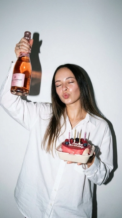 Ультрареалистичная фотография в эстетике Party Girl / Birthday Aesthetic, стилизация под случайный снимок на компактную цифровую камеру начала 2000-х.
 девушка празднует день рождения. На ней свободная белая рубашка оверсайз на пуговицах. В одной руке она высоко держит бутылку розового шампанского, в другой — небольшой бенто-торт со светлыми свечами и свежими ягодами.

Глаза закрыты, на лице игривое выражение с лёгким «поцелуйным» жестом губ. Не меняй прическу модели.

Съёмка со вспышкой прямо в камеру на фоне простой молочно-белой стены. Жёсткий свет создаёт характерную чёткую тень за фигурой, яркие блики на коже, волосах, бутылке и глазури торта. Атмосфера спонтанного домашнего праздничного снимка: немного несовершенная композиция, лёгкая зернистость, естественная текстура кожи, реалистичные отражения и мелкие детали.

Стиль: 2000s digital camera, lo-fi party aesthetic, candid snapshot, hard flash, authentic lifestyle photography, natural skin texture, realistic colors, high detail, 4K.

Без: студийной постановки, мягкого идеального света, пластиковой кожи, чрезмерной ретуши, CGI, мультяшности, искажённых рук и пальцев, лишних пальцев, деформированной бутылки или торта, артефактов текста, логотипов и водяных знаков.