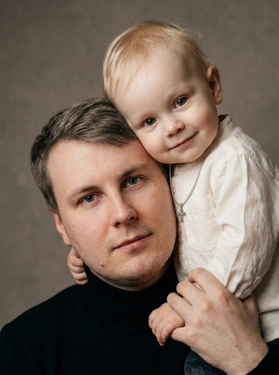 Родитель и ребёнок с внешностью как на приложенных фото, художественная семейная студийная фотосессия. Композиция — вертикальный портрет, крупный план лиц. Родитель внизу смотрит прямо в камеру, лицо фронтально. Ребёнок сверху, голова повернута набок, щека лежит на голове родителя, смотрит в камеру с нежной улыбкой. Рука ребёнка обнимает родителя за шею, видна в кадре, родитель держит руку ребёнка своей рукой.
Одежда: ребёнок в белой льняной рубашке, родитель в чёрной водолазке. 
Фон — тёмно-серый почти чёрный однотонный студийный, создаёт контраст. Освещение — мягкое боковое студийное, создаёт объём и глубину.
Чёрно-белая художественная фотография с выразительными контрастами. Атмосфера нежная, интимная, спокойная.