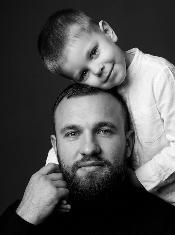 Родитель и ребёнок с внешностью как на приложенных фото, художественная семейная студийная фотосессия. Композиция — вертикальный портрет, крупный план лиц. Родитель внизу смотрит прямо в камеру, лицо фронтально. Ребёнок сверху, голова повернута набок, щека лежит на голове родителя, смотрит в камеру с нежной улыбкой. Рука ребёнка обнимает родителя за шею, видна в кадре, родитель держит руку ребёнка своей рукой.
Одежда: ребёнок в белой льняной рубашке, родитель в чёрной водолазке. 
Фон — тёмно-серый почти чёрный однотонный студийный, создаёт контраст. Освещение — мягкое боковое студийное, создаёт объём и глубину.
Чёрно-белая художественная фотография с выразительными контрастами. Атмосфера нежная, интимная, спокойная.