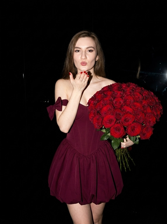 Сохранить внешность на 100%. Черты лица не изменять.
Ultra-realistic vertical 9:16 photo, Y2K luxury nightlife, paparazzi flash.

Полная темнота.
Фон почти полностью чёрный — ресторан не читается, только намёки: слабые отражения, едва заметные силуэты, глянец поверхностей. Всё пространство «съедено» тьмой.

Единственный источник света — жёсткая вспышка камеры, бьёт прямо в лицо и фигуру, создаёт резкий контраст, глубокие тени, эффект ночного paparazzi-кадра начала 2000-х.

Женщина в платье:
— глубокий винно-бордовый / dark cherry цвет
— корсетное мини без бретелей
— жёсткий sculptural corset
— короткая объёмная bubble-юбка
— off-shoulder банты
— плотная дорогая couture-ткань
— силуэт 1:1, без изменений

В руках — огромный, сверхпышный букет цветов, почти перекрывает корпус:
— тяжёлый, статусный,
— максимально плотный,
— luxury florist look.

Действие:
— она посылает воздушный поцелуй в камеру,
— взгляд дерзкий, холодный, уверенный,
— без улыбки.

Эффект:
— вспышка «вырывает» её из темноты,
— кожа и ткань ловят свет,
— фон проваливается в чёрный.

Стиль:
Y2K rich bitch, flash-only lighting, dark luxury, paparazzi moment, ultra-realistic, expensive, iconic, no softness, no romance, pure power.

Какие будут цветы: В руках у нее букет тёмно-красных роз.