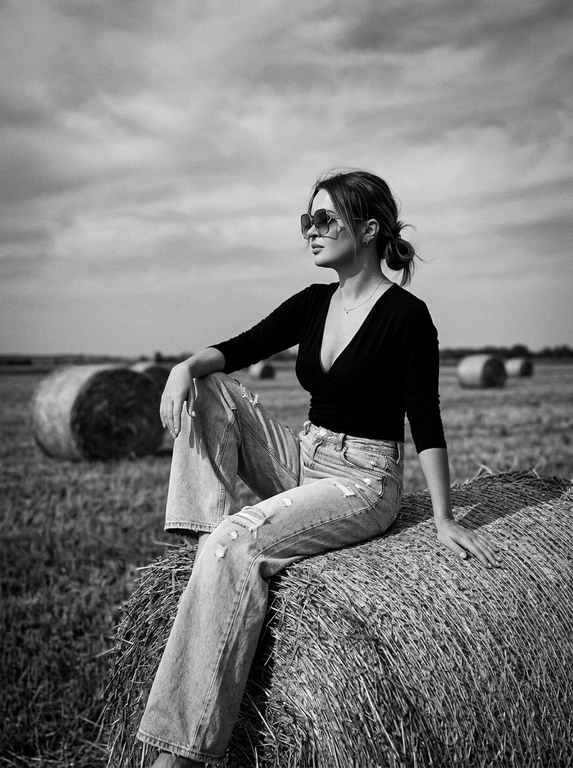 Чёрно-белая high-fashion editorial фотография в сельской местности, женщина с внешностью как на референсе, черты лица не изменять, композиция — более общий план (wide shot), камера на среднем расстоянии, фигура полностью в кадре с окружающим пространством, она сидит на большом тюке сена в поле, корпус слегка развернут, одна нога свободно опущена вниз, вторая согнута, создавая расслабленную, но выразительную позу, руки естественно лежат на колене и на поверхности тюка, взгляд направлен в сторону, профиль, выражение спокойное и уверенное, одежда: чёрный топ с V-образным вырезом и объёмные светлые широкие джинсы с потёртостями и рваными элементами, аксессуары: крупные солнцезащитные очки, минималистичные украшения, фон: открытое поле с несколькими тюками сена на разной глубине, создающими перспективу, линия горизонта ниже центра кадра, чтобы подчеркнуть небо, освещение: жёсткий естественный дневной свет с мягкими облаками, формирует выразительные тени и контраст, стиль: кинематографический, плёночное зерно, высокая детализация, реалистичная текстура кожи и ткани, редакционный fashion стиль, акцент на атмосфере и пространстве кадра


Какая будет прическа?: НЕ изменять мою прическу