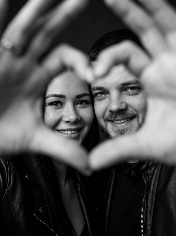 Ultra-realistic editorial photography of a smiling couple in a romantic and cinematic composition. The couple is centered in the background of the image, looking directly at the camera with a spontaneous, happy, and natural expression. In the foreground, extremely close to the lens and slightly out of focus, the couple’s hands form a heart shape perfectly framing their faces.

The composition creates strong visual depth, with sharp focus on the eyes and faces inside the heart formed by the hands. The hands occupy a large portion of the foreground, artistically blurred around the edges, creating a sophisticated cinematic effect.

Intimate, emotional, and elegant aesthetic inspired by modern perfume campaigns and premium lifestyle editorials. Soft studio lighting with delicate contrast and natural shadows. Minimalistic dark blurred background.

The couple wears minimalistic black leather jackets and sophisticated dark clothing. Genuine expressions, strong emotional connection, contemporary romantic atmosphere. Hyper-realistic skin texture, visible pores, detailed hair strands, authentic human appearance.

Black and white photography, rich tones, deep blacks, and soft highlights. 85mm lens, shallow depth of field, cinematic portrait photography, luxury editorial style, ultra detailed, hyper realistic, 8K.