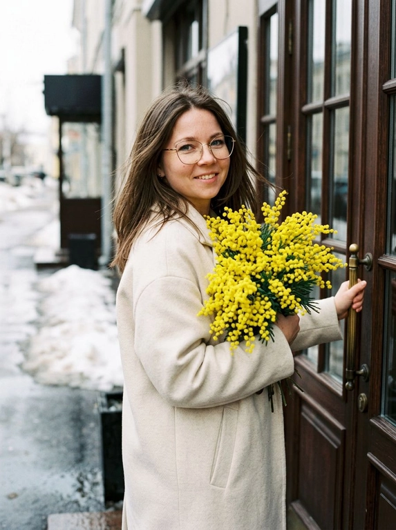 Сохранить черты лица и внешность, возраст, натуральный тон без ретуши. Средне-крупный план (от талии вверх), камера слева от женщины, лицо занимает не менее 35% кадра. Женщина стоит у входа в городское кафе с деревянной дверью и стеклянными вставками, корпус слегка развернут в 3/4 к камере. Лицом к двери, одной рукой держит ручку закрытой двери, другой прижимает к груди огромный пышный букет ярко-жёлтой мимозы. На ней элегантное светлое пальто (молочный оттенок), свободный крой. Волосы как на исходном фото слегка взъерошены ветром Мягкое выражение лица, лёгкая улыбка, взгляд в камеру. Атмосфера начала весны: тающий снег по краям улицы, влажный асфальт, мягкий рассеянный дневной свет, лёгкая прохлада в воздухе. Цветовая палитра нейтральная с ярким акцентом жёлтой мимозы. Фотореализм, натуральная текстура ткани и цветов, без HDR, без пластика, мягкий контраст, 35mm look.