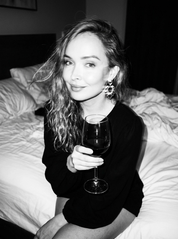 Create a portrait, transfer the face from the reference photo without changing facial features, proportions, or expression, preserve full identity. Aesthetic black-and-white nighttime Y2K photograph captured with direct on-camera flash, shot in motion.
A woman is sitting on the edge of a white, slightly rumpled bed, the frame taken from a slightly elevated angle. She wears a stylish black top with voluminous sleeves and large round statement earrings. In one hand she holds an elegant glass of dark wine. Her body is subtly moving, creating natural motion blur across the face and shoulders, as if the moment was caught mid-gesture. The gaze is languid and directed toward the camera, with a soft, intimate smile. Hair falls in large waves with loose strands drifting across the face, slightly displaced by movement. Lighting is harsh flash against deep night shadows, high contrast with blown highlights and strong falloff, visible film grain. Skin texture remains real and alive, no smoothing, enhanced by flash glare. The mood is high-fashion editorial, Parisian chic, old-money sensuality, nighttime melancholy, spontaneous and cinematic, shot in a raw 35mm film aesthetic with pronounced motion blur and lived-in realism.