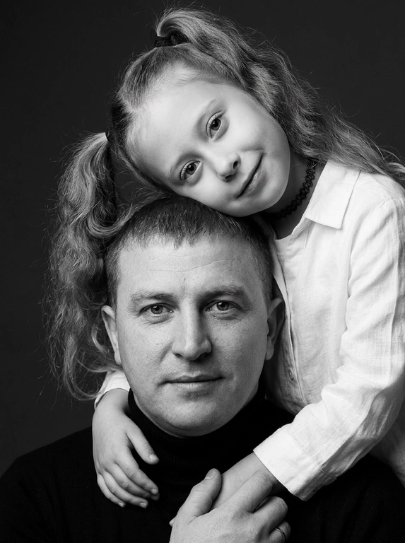 Родитель и ребёнок с внешностью как на приложенных фото, художественная семейная студийная фотосессия. Композиция — вертикальный портрет, крупный план лиц. Родитель внизу смотрит прямо в камеру, лицо фронтально. Ребёнок сверху, голова повернута набок, щека лежит на голове родителя, смотрит в камеру с нежной улыбкой. Рука ребёнка обнимает родителя за шею, видна в кадре, родитель держит руку ребёнка своей рукой.
Одежда: ребёнок в белой льняной рубашке, родитель в чёрной водолазке. 
Фон — тёмно-серый почти чёрный однотонный студийный, создаёт контраст. Освещение — мягкое боковое студийное, создаёт объём и глубину.
Чёрно-белая художественная фотография с выразительными контрастами. Атмосфера нежная, интимная, спокойная.