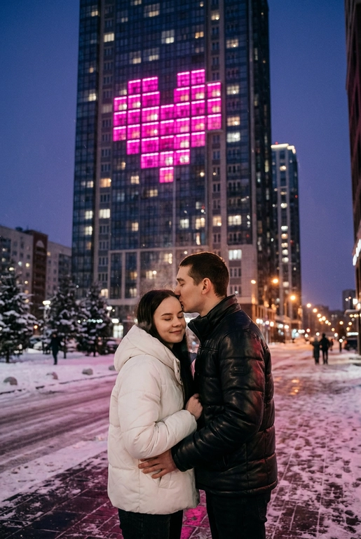 Создай не меняя черты лица. Романтический снимок пары, стоящей на заснеженной городской улице поздним вечером. Главный объект – крупный план пары: мужчина в черной кожаной дутой зимней куртке нежно целует женщину в белой дутой куртке в лоб, она стоит с прикрытыми глазами, улыбаясь. За ними возвышается массивный современный жилой небоскреб, на фасаде которого окна на нескольких этажах подсвечены ярким  светом, формируя огромное пиксельное сердце. Освещение драматичное, контрастное, с преобладанием холодных тонов в небе и тёплого, неонового розового свечения от здания. Снег на земле отражает свет. Урбанистическая, зимняя, кинематографическая фотография, атмосферный кадр.