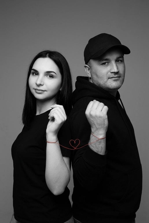Фотореалистичный чёрно-белый студийный портрет пары, стоящей спина к спине, слегка повернувшись к камере. Оба в минималистичных чёрных образах (худи и футболка), с мягкими уверенными выражениями лица, естественный внешний вид. Тонкая красная нить, связанная вокруг их запястий, соединяет их и образует маленькое сердце между ними. Чистый светло-серый студийный фон, мягкое рассеянное освещение, высокая детализация текстуры кожи, резкий фокус, кинематографичная композиция, вертикальный формат 9:16, минималистичная эстетика, высокое разрешение, профессиональная фотография.

Доп. характеристики: У девушки Цвет волос черный